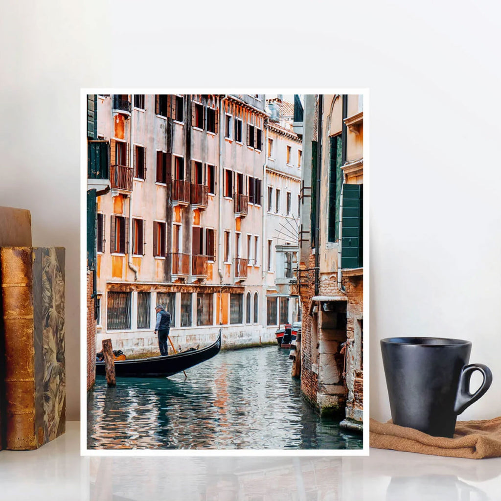 FRAMED VENICE CANAL ART PRINT FEATURING A GONDOLA AND HISTORIC BUILDINGS, DISPLAYED ON A WHITE SURFACE BESIDE A BLACK MUG AND BOOKS.