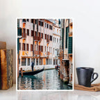 FRAMED VENICE CANAL ART PRINT FEATURING A GONDOLA AND HISTORIC BUILDINGS, DISPLAYED ON A WHITE SURFACE BESIDE A BLACK MUG AND BOOKS.