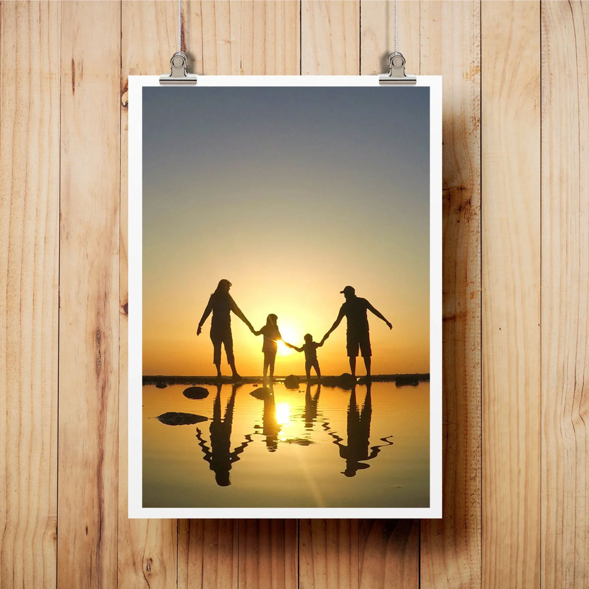 FAMILY SILHOUETTE ART PRINT FEATURING A FAMILY HOLDING HANDS IN SHALLOW WATER AT SUNSET, DISPLAYED ON A LIGHT WOOD WALL WITH METAL CLIPS.