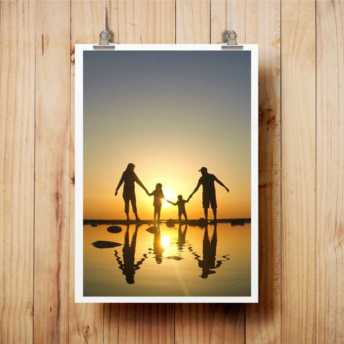 FAMILY SILHOUETTE ART PRINT FEATURING A FAMILY HOLDING HANDS IN SHALLOW WATER AT SUNSET, DISPLAYED ON A LIGHT WOOD WALL WITH METAL CLIPS.