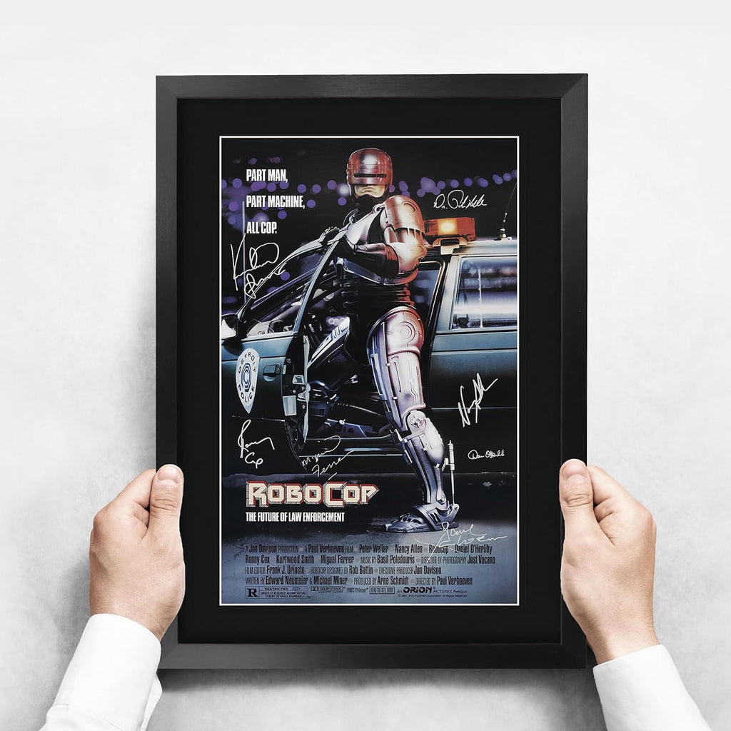 FRAMED ROBOCOP MOVIE POSTER FEATURING ROBOCOP ON A MOTORCYCLE WITH A POLICE CAR, HELD BY TWO HANDS AGAINST A LIGHT GRAY WALL.