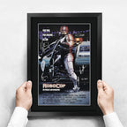 FRAMED ROBOCOP MOVIE POSTER FEATURING ROBOCOP ON A MOTORCYCLE WITH A POLICE CAR, HELD BY TWO HANDS AGAINST A LIGHT GRAY WALL.