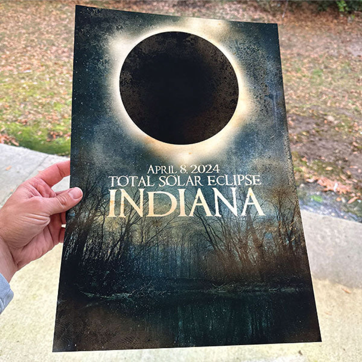 HAND HOLDING A PRINTED TOTAL SOLAR ECLIPSE POSTER FEATURING THE APRIL 8, 2024 INDIANA ECLIPSE OVER A DARK FOREST LANDSCAPE.