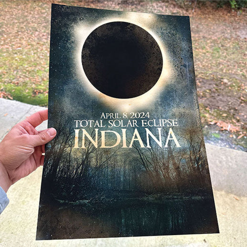 HAND HOLDING A PRINTED TOTAL SOLAR ECLIPSE POSTER FEATURING THE APRIL 8, 2024 INDIANA ECLIPSE OVER A DARK FOREST LANDSCAPE.