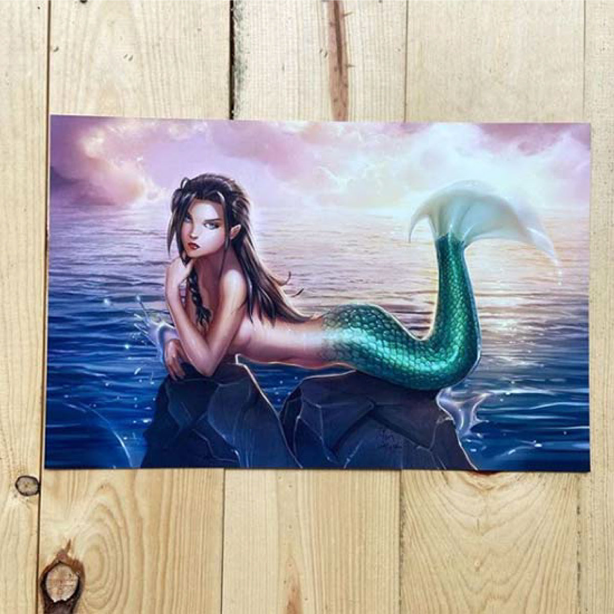 RECTANGULAR ART PRINT FEATURING A MERMAID WITH A GREEN TAIL RESTING ON AN OCEAN ROCK BENEATH A PINK SKY, DISPLAYED ON A LIGHT WOOD SURFACE.