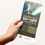 HAND HOLDING A CUSTOM PRINTED PROMOTIONAL POSTCARD FEATURING A WINTER CLEARANCE SALE AND OUTDOOR CAMPING DESIGN ON A CLEAN WHITE BACKGROUND.