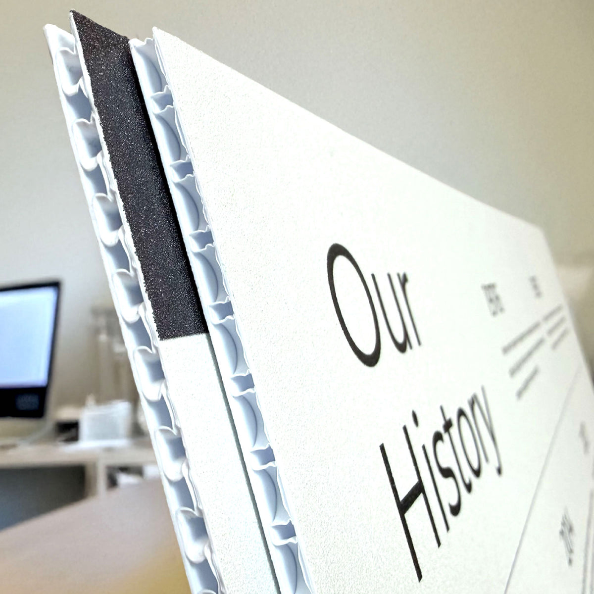 CLOSE-UP OF A CUSTOM PRINTED BUBBLE CORE PLASTIC SIGN FEATURING “OUR HISTORY” TEXT, SHOWING THE LIGHTWEIGHT LAYERED CORE AND PRINTED SURFACE IN AN INDOOR OFFICE SETTING.