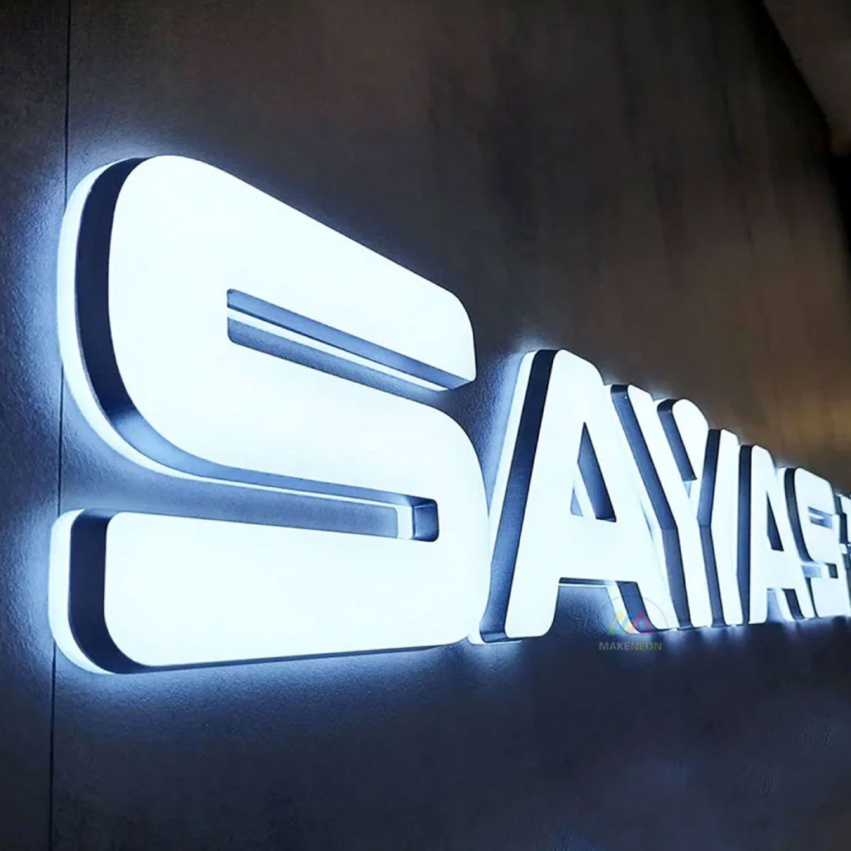 CLOSE-UP OF LARGE 3D ILLUMINATED LETTERS SPELLING “SAYAS” WITH BRIGHT WHITE LED LIGHTING, MOUNTED ON A DARK TEXTURED WALL.