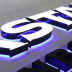 3D ILLUMINATED LETTERS WITH WHITE FACES, BLACK RETURNS, AND BLUE-PURPLE LED HALO LIGHTING, DISPLAYED ON A LIGHT GRAY BACKGROUND.