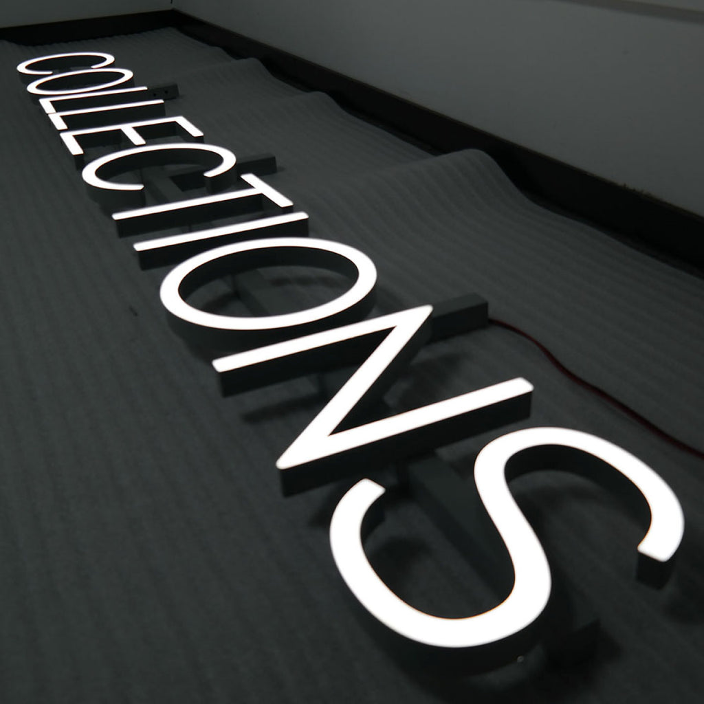 3D ILLUMINATED LETTERS SPELLING “COLLECTIONS” WITH BRIGHT WHITE LED LIGHTING, MOUNTED ON A DARK TEXTURED WALL.
