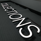 3D ILLUMINATED LETTERS SPELLING “COLLECTIONS” WITH BRIGHT WHITE LED LIGHTING, MOUNTED ON A DARK TEXTURED WALL.