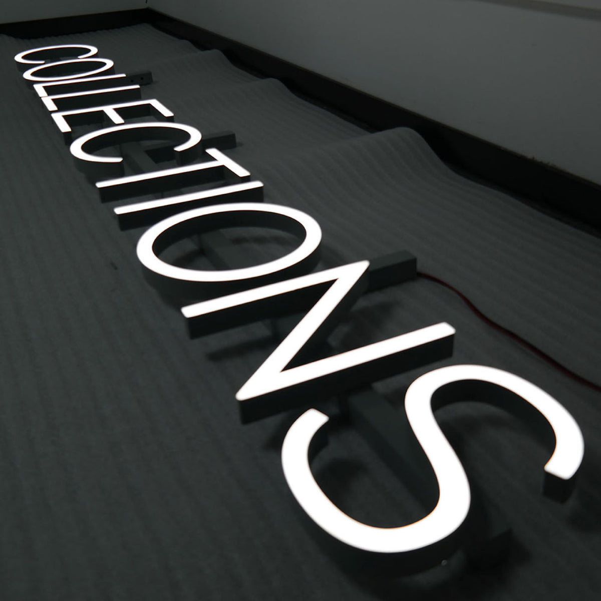 3D ILLUMINATED LETTERS SPELLING “COLLECTIONS” WITH BRIGHT WHITE LED LIGHTING, MOUNTED ON A DARK TEXTURED WALL.