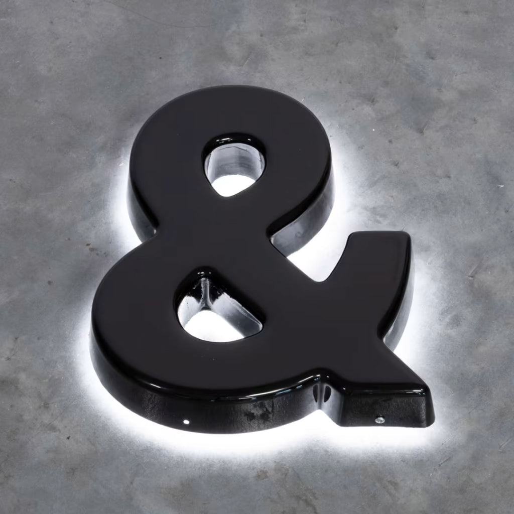 GLOSSY BLACK 3D AMPERSAND SIGN WITH WHITE LED HALO BACKLIGHTING, MOUNTED ON A TEXTURED GRAY CONCRETE WALL.
