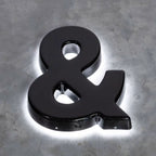 GLOSSY BLACK 3D AMPERSAND SIGN WITH WHITE LED HALO BACKLIGHTING, MOUNTED ON A TEXTURED GRAY CONCRETE WALL.