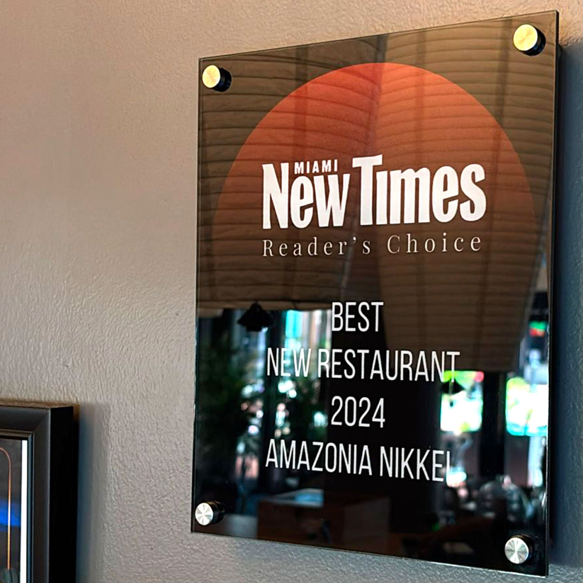 CUSTOM PRINTED GLOSSY BLACK ACRYLIC WALL PLAQUE FEATURING THE MIAMI NEW TIMES READER’S CHOICE BEST NEW RESTAURANT 2024 AWARD FOR AMAZONIA NIKKEI, MOUNTED ON A BEIGE INTERIOR WALL.