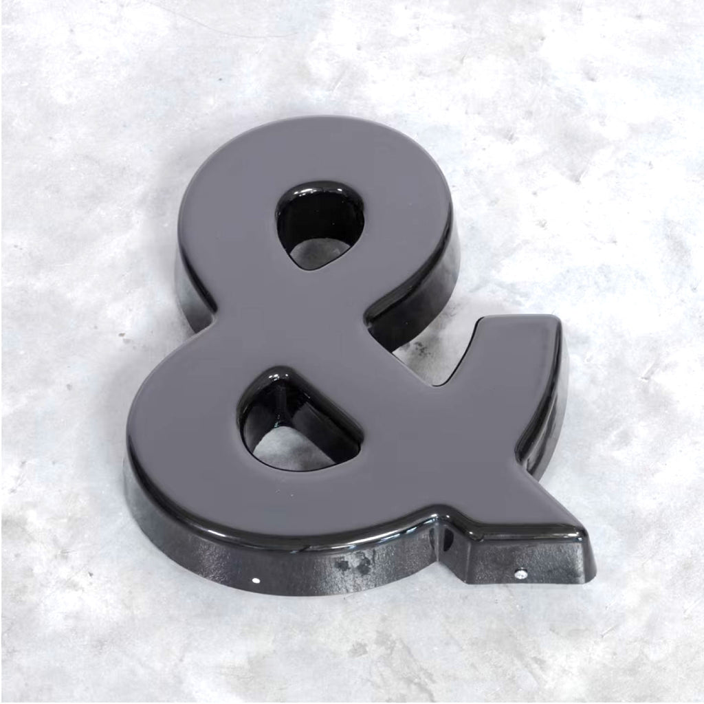 GLOSSY BLACK 3D AMPERSAND SIGN WITH A DIMENSIONAL FINISH, DISPLAYED AT AN ANGLE ON A TEXTURED LIGHT GRAY SURFACE.