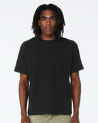 A man with long black dreadlocks wearing a plain black short-sleeve crewneck t-shirt and light olive pants, standing against a solid white studio background.