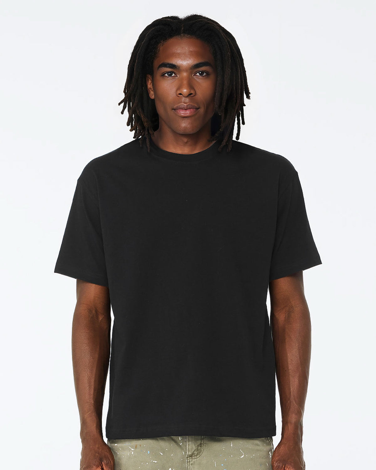 A man with long black dreadlocks wearing a plain black short-sleeve crewneck t-shirt and light olive pants, standing against a solid white studio background.
