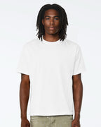A man with dark dreadlocks wearing a plain white short-sleeve crewneck t-shirt and light olive pants, standing against a solid white studio background.