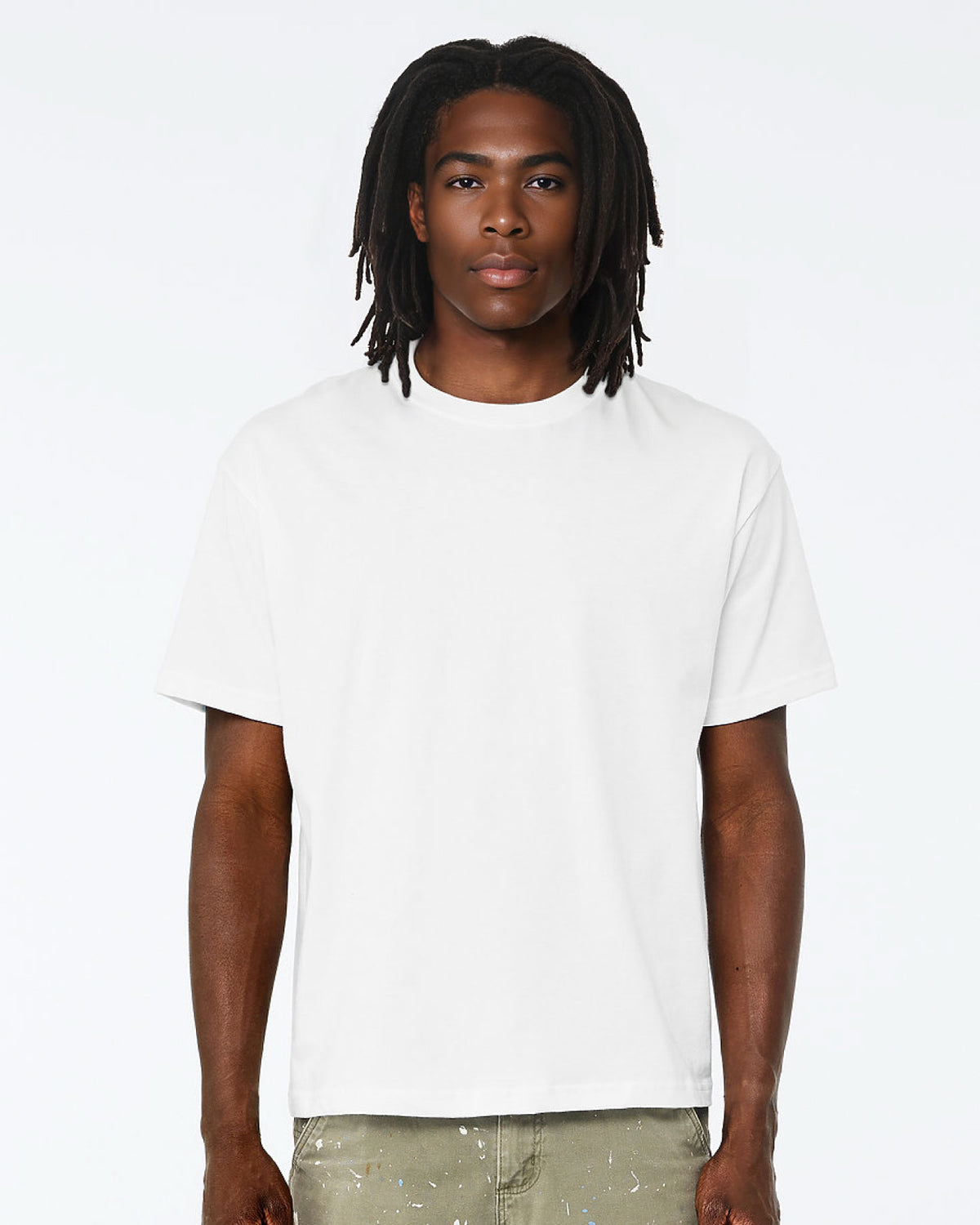 A man with dark dreadlocks wearing a plain white short-sleeve crewneck t-shirt and light olive pants, standing against a solid white studio background.