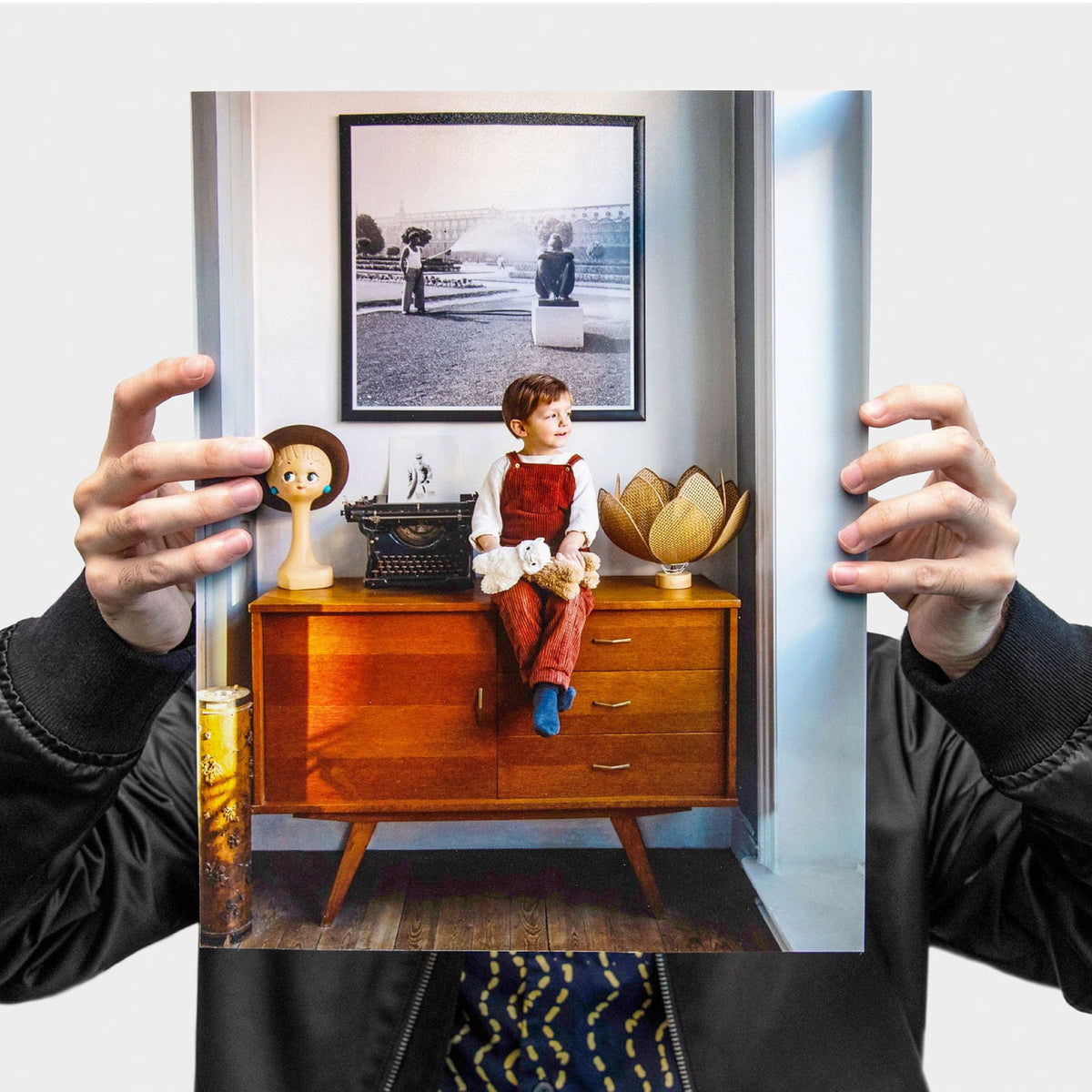 PERSON HOLDING A LARGE FULL-COLOR PRINTED POSTER FEATURING A CHILD SITTING ON A WOODEN CABINET IN A STYLISH INTERIOR SETTING.
