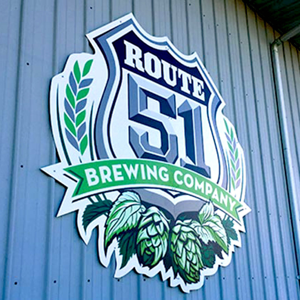 CUSTOM PRINTED ALUMINUM SIGN FEATURING ROUTE 51 BREWING COMPANY BRANDING WITH A SHIELD, WHEAT, HOPS, AND GREEN BANNER GRAPHICS, MOUNTED ON A BLUE CORRUGATED METAL EXTERIOR WALL.