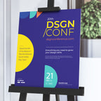 CUSTOM PRINTED FOAM BOARD POSTER FEATURING DSGN / CONF CONFERENCE GRAPHICS, DISPLAYED ON A BLACK EASEL STAND IN A MODERN INDOOR EVENT SETTING.