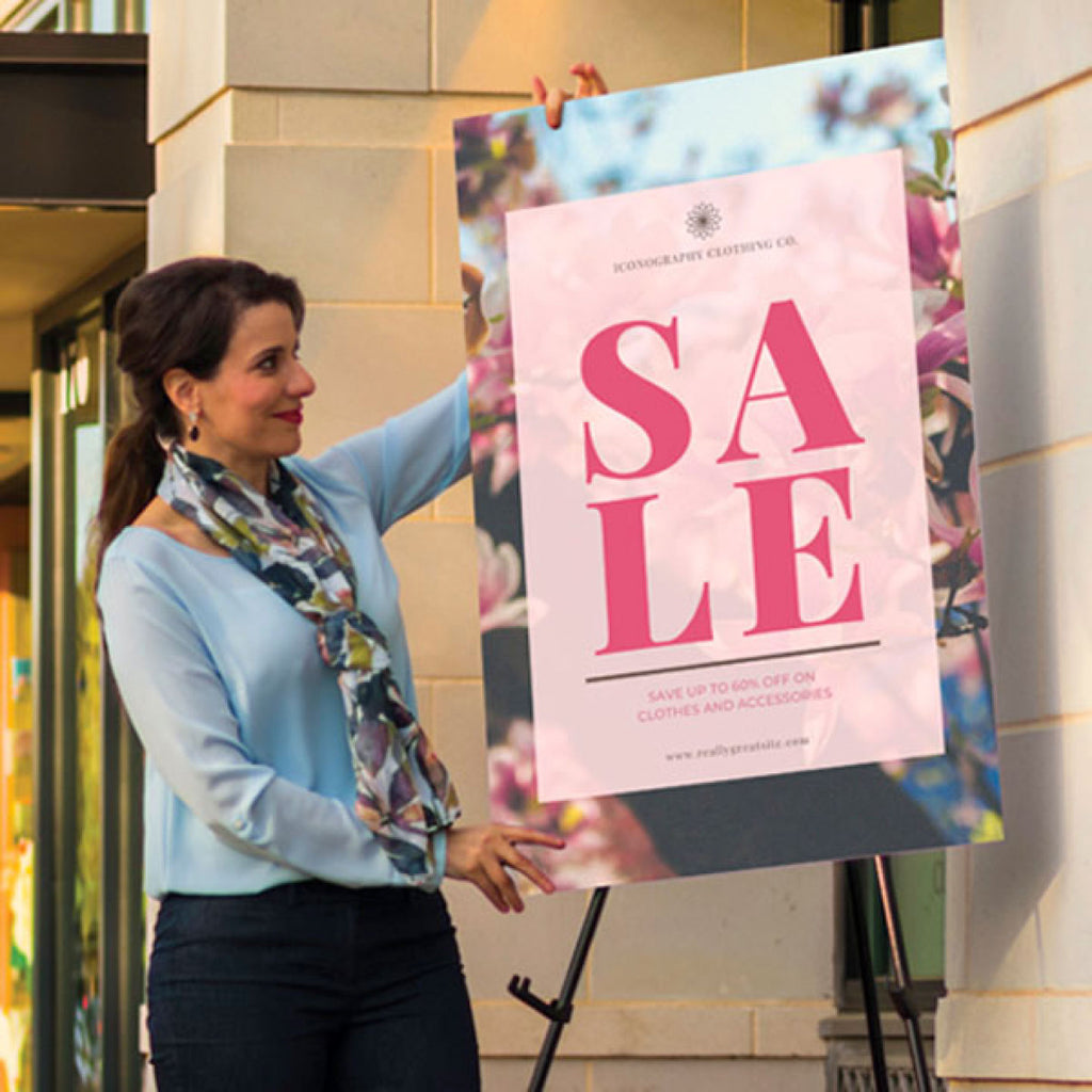 CUSTOM PRINTED FOAM BOARD SIGN FEATURING A LARGE “SALE” PROMOTION WITH FLORAL GRAPHICS, DISPLAYED ON AN EASEL AT A BUILDING ENTRANCE.
