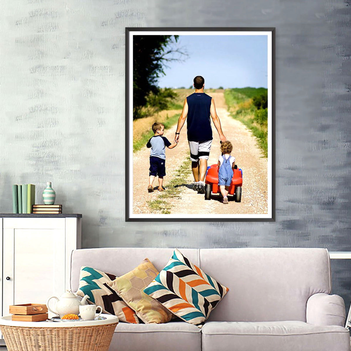 CUSTOM PRINTED PAPER POSTER FEATURING A FAMILY PHOTOGRAPH OF A FATHER AND TWO CHILDREN WALKING ON A DIRT ROAD, FRAMED AND DISPLAYED ON A TEXTURED GRAY WALL ABOVE A SOFA.