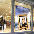 LARGE CUSTOM PRINTED STYRENE RETAIL DISPLAY SIGN FEATURING A SUMMER SWIMWEAR SALE PROMOTION WITH VIBRANT GRAPHICS, DISPLAYED IN A STOREFRONT WINDOW BESIDE MANNEQUINS AND CLOTHING RACKS.