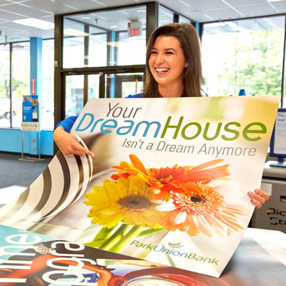 CUSTOM PRINTED STYRENE SIGN FEATURING A PARK UNION BANK HOME PROMOTION WITH COLORFUL FLORAL GRAPHICS, HELD BY A WOMAN IN A BANK LOBBY.