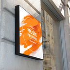 CUSTOM PRINTED TRANSLUCENT BACKLIT SIGN INSERT FEATURING A BOLD ORANGE AND WHITE PRESENT MUSIC FESTIVAL POSTER, DISPLAYED IN A FRAMED SIGN MOUNTED ON A GRAY STONE WALL.