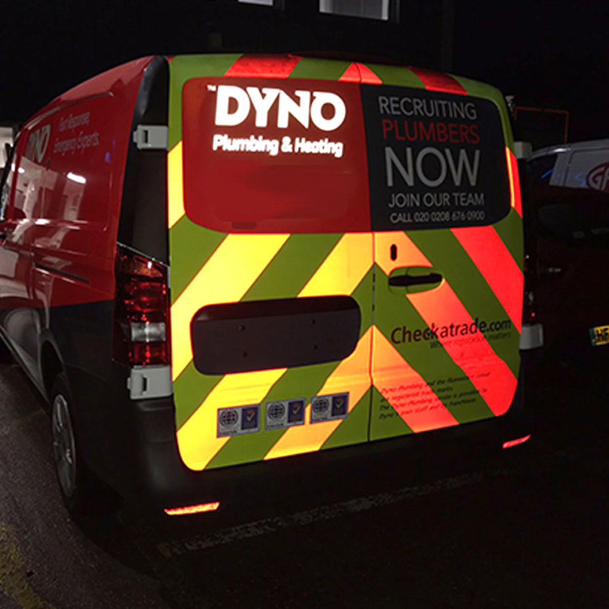 COMMERCIAL VAN AT NIGHT FEATURING BRIGHT REFLECTIVE VINYL GRAPHICS WITH RED, YELLOW, AND GREEN DIAGONAL STRIPES, DYNO PLUMBING & HEATING BRANDING, AND RECRUITMENT MESSAGING FOR HIGH VISIBILITY IN LOW-LIGHT CONDITIONS.
