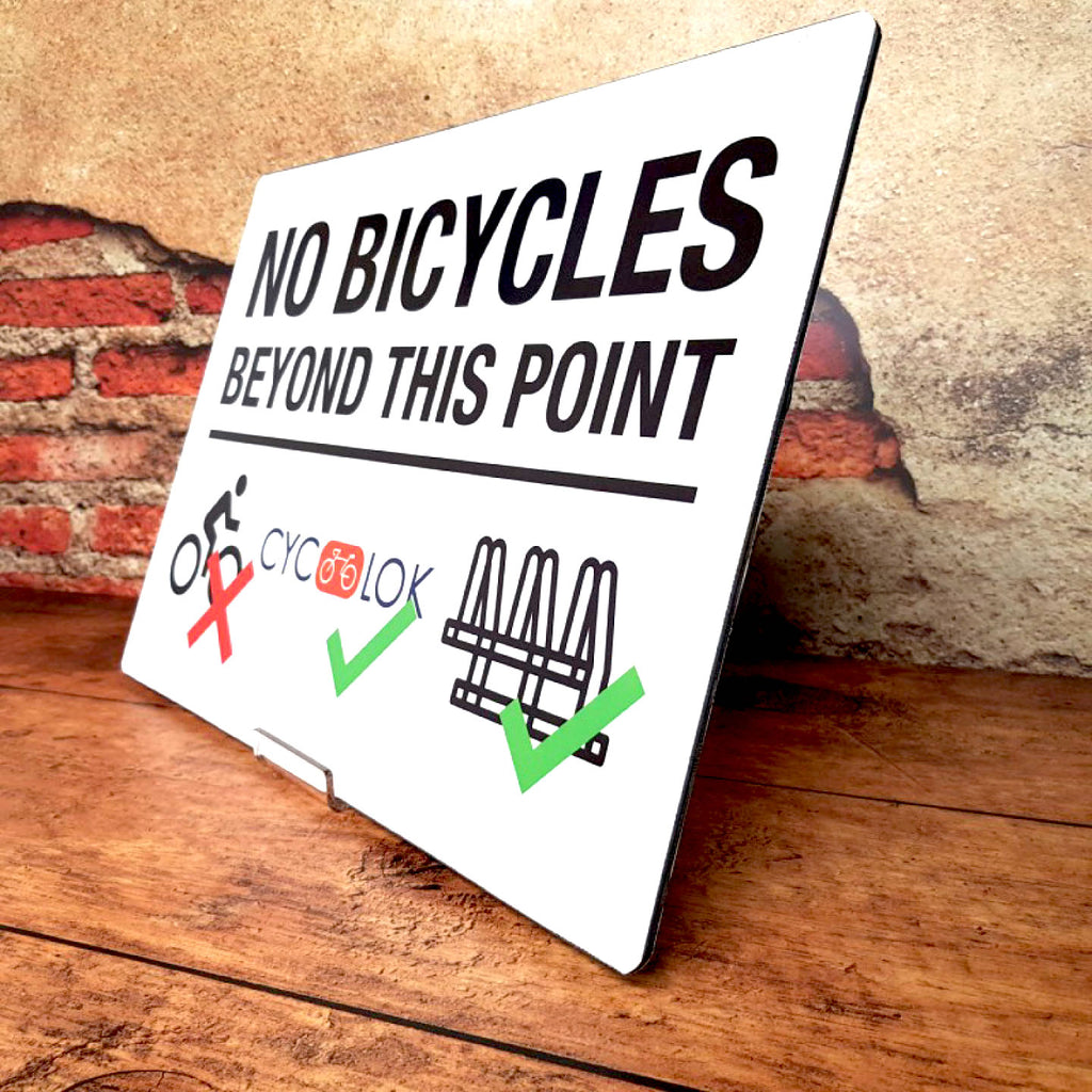 CUSTOM PRINTED ACM WARNING SIGN FEATURING A “NO BICYCLES BEYOND THIS POINT” MESSAGE WITH A CROSSED-OUT BICYCLE SYMBOL AND DIRECTIONAL GRAPHICS, DISPLAYED AGAINST AN EXTERIOR BRICK AND STUCCO WALL.
